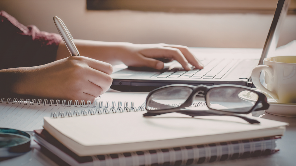 hands writing in notebook with glasses nearby.