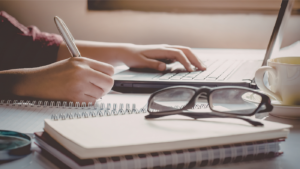 hands writing in notebook with glasses nearby.