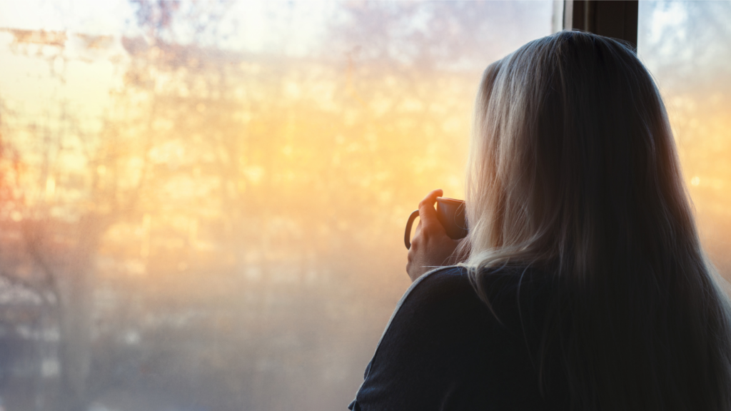 Woman holding a mug by a window in soft morning light, reflecting gentle wellness habits for women with low energy.